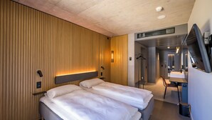Habitació d'hotel moderna amb dos llits, un mur de panells de fusta, televisió integrada i un bany amb mirall i banyera.