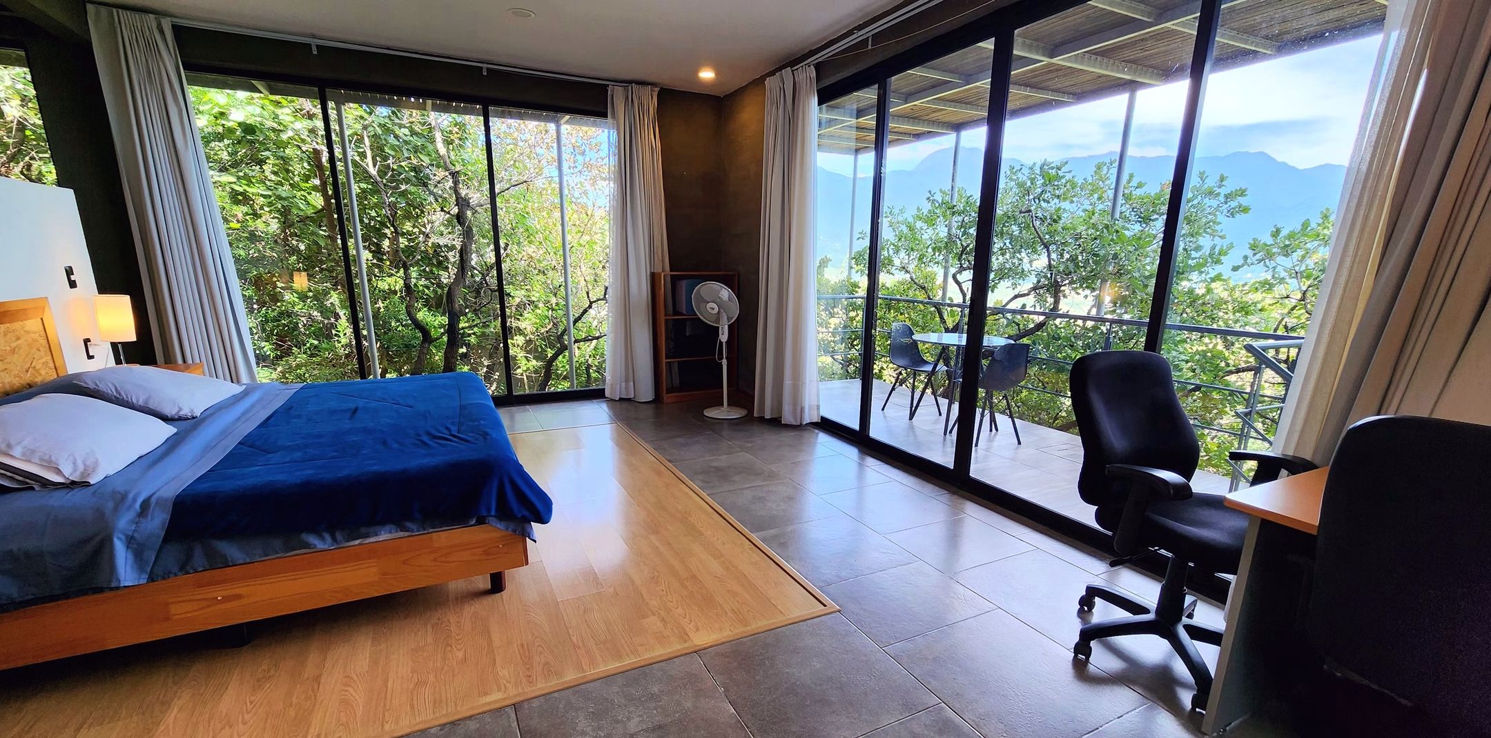 A bedroom with a bed, a desk, and a chair. There is a large window with a view of trees and mountains.