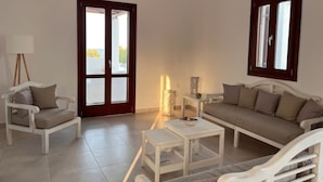 Ein Wohnzimmer mit Sofa, Sessel, Couchtisch und Lampe. Zwei Türen und Fenster mit Vorhängen sind zu sehen.