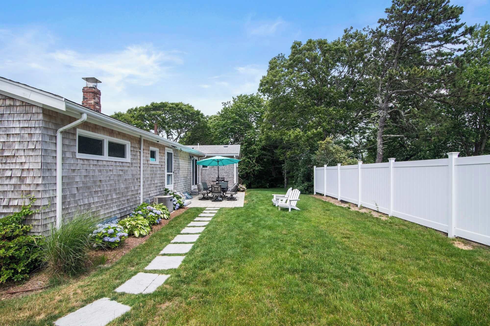 A residential backyard with a house, a patio with outdoor furniture, a white fence, and a lawn.