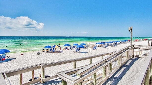 A beach with a wooden boardwalk, blue umbrellas, and beach chairs.