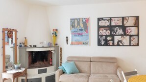 A living room with a beige sofa, a small table with a lamp, a fireplace, and a wall with framed photos and a poster. - Single-family home (Scandicci)