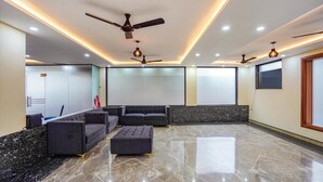 A spacious living area with a sectional sofa, ottoman, ceiling fans, recessed lighting, and a marble floor. - The Grand Mansion Hotel (Greater Noida)
