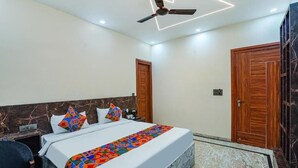 A hotel room with a bed, a desk, a chair, a ceiling fan, and wooden doors. - The Grand Mansion Hotel (Greater Noida)
