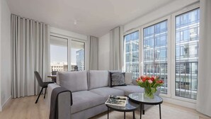 Una sala moderna con un sofá gris, una mesa redonda de centro y vistas a un paisaje urbano a través de amplios ventanales.