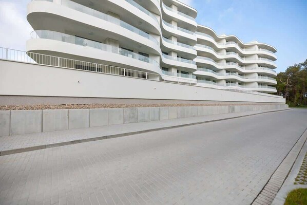 Un edificio moderno de varios pisos con balcones, un sendero pavimentado y cielo despejado.