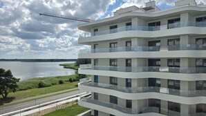 A multi-story building with balconies, a view of a lake, and a construction crane in the background. - Stylish Apartment | Parking, Balcony, Close to the Beach (Rogowo)