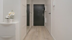 Un pasillo estrecho con una puerta oscura al final, paredes blancas y un estante pequeño con una flor.
