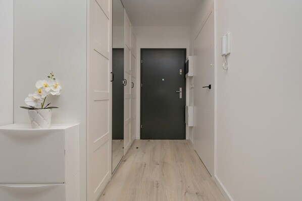 A narrow hallway with a dark door at the end, white walls, and a small white shelf with a flower.