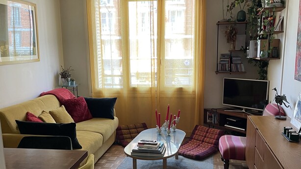 Una sala de estar con un sofá, una mesa de centro y un televisor. Hay ventanas con cortinas, una estantería y una lámpara colgante.