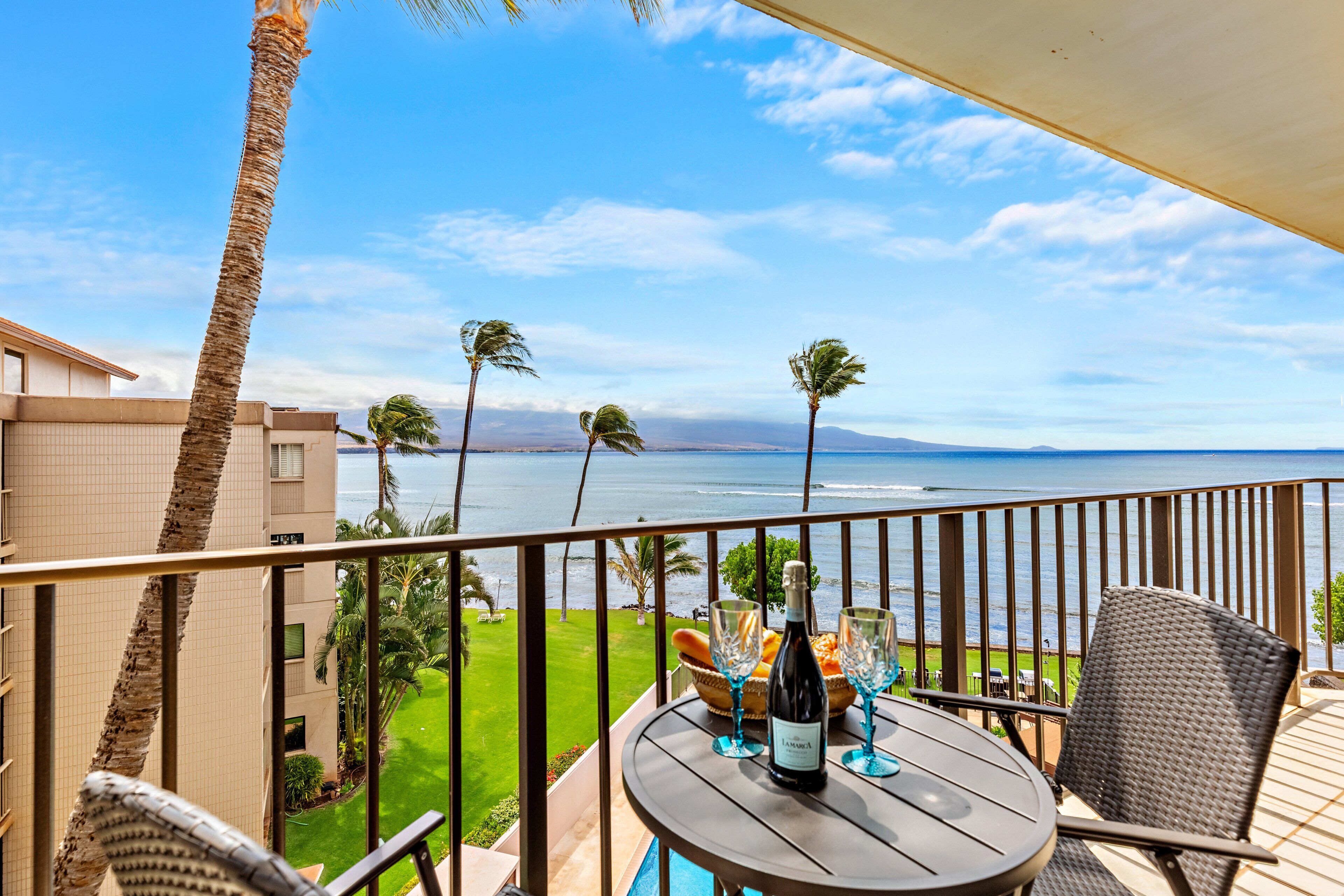 A balcony with a table set for two, overlooking a coastal view with palm trees and a clear sky.