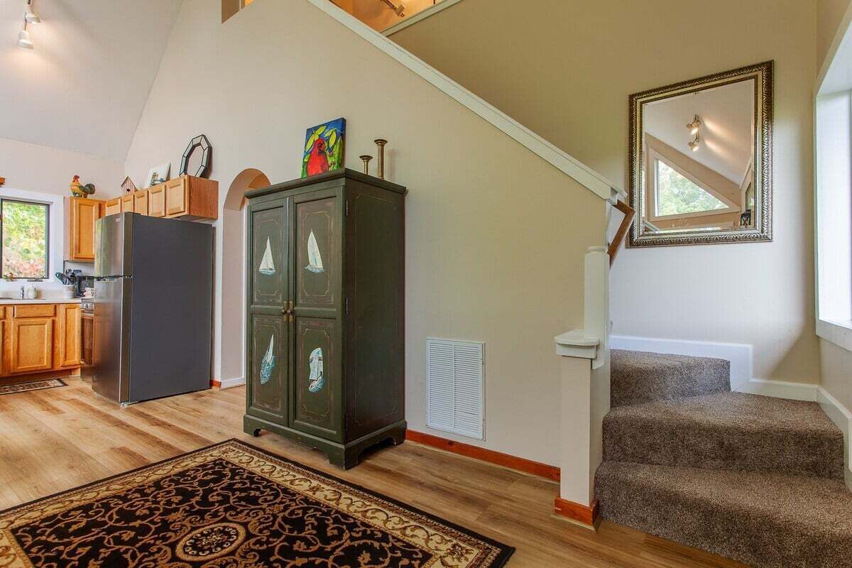 Una cocina amplia con armarios de madera, un refrigerador y un mueble oscuro con objetos decorativos. Una escalera con peldaños alfombrados lleva a un rellano con un espejo.