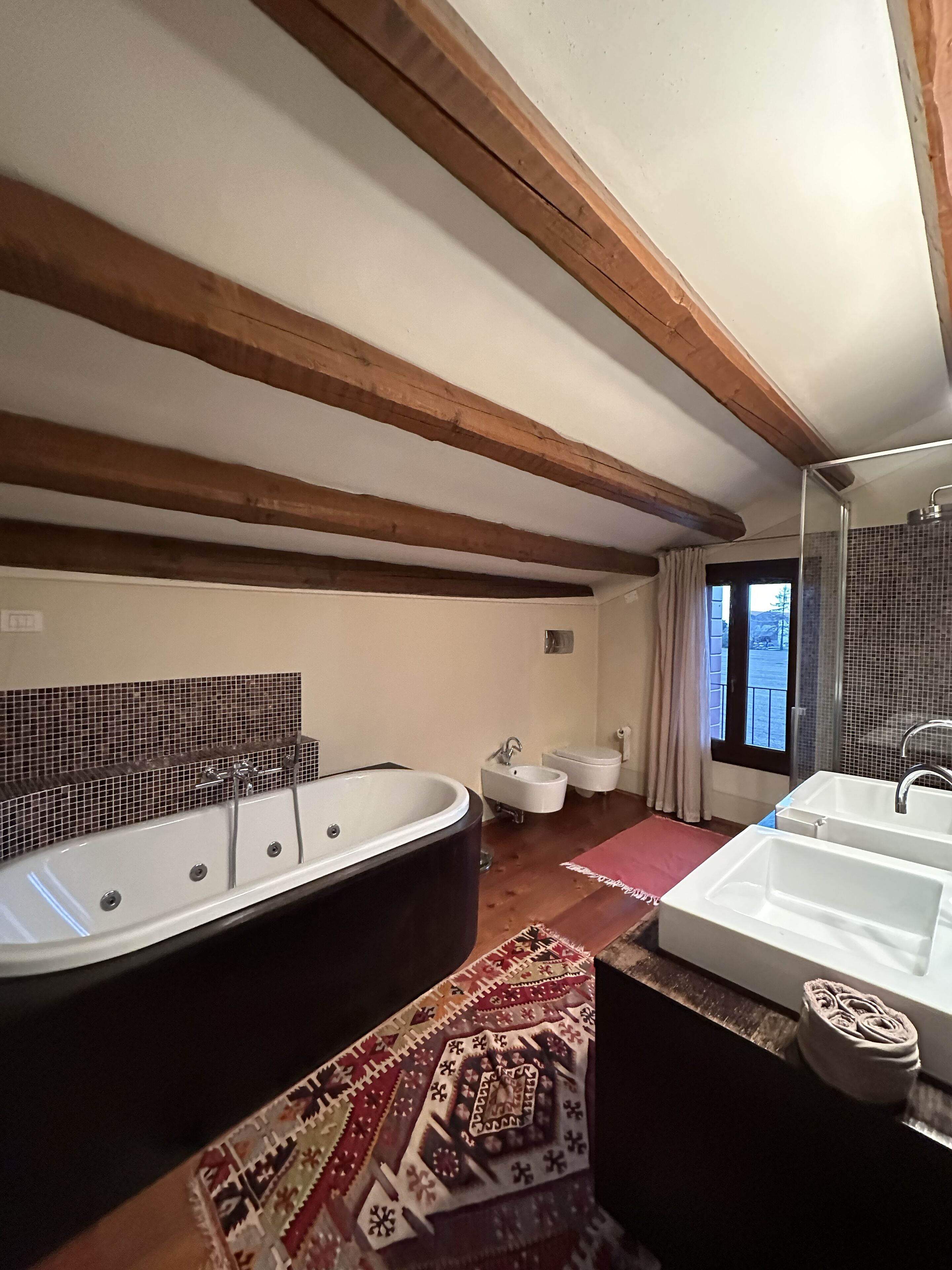 A bathroom with a large freestanding bathtub, a double sink, and a bidet. The room has wooden beams on the ceiling and a view of the outdoors through a window.