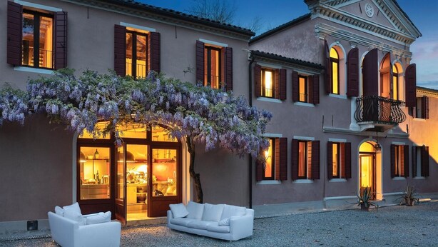 A building with a large glass door entrance, a covered outdoor seating area with white sofas, and a pergola with purple flowers.