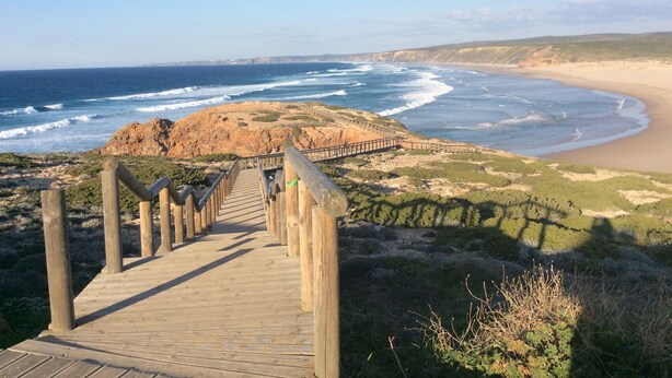 Une promenade en bois menant à une plage, avec un promontoire rocheux et une vue sur l’océan.