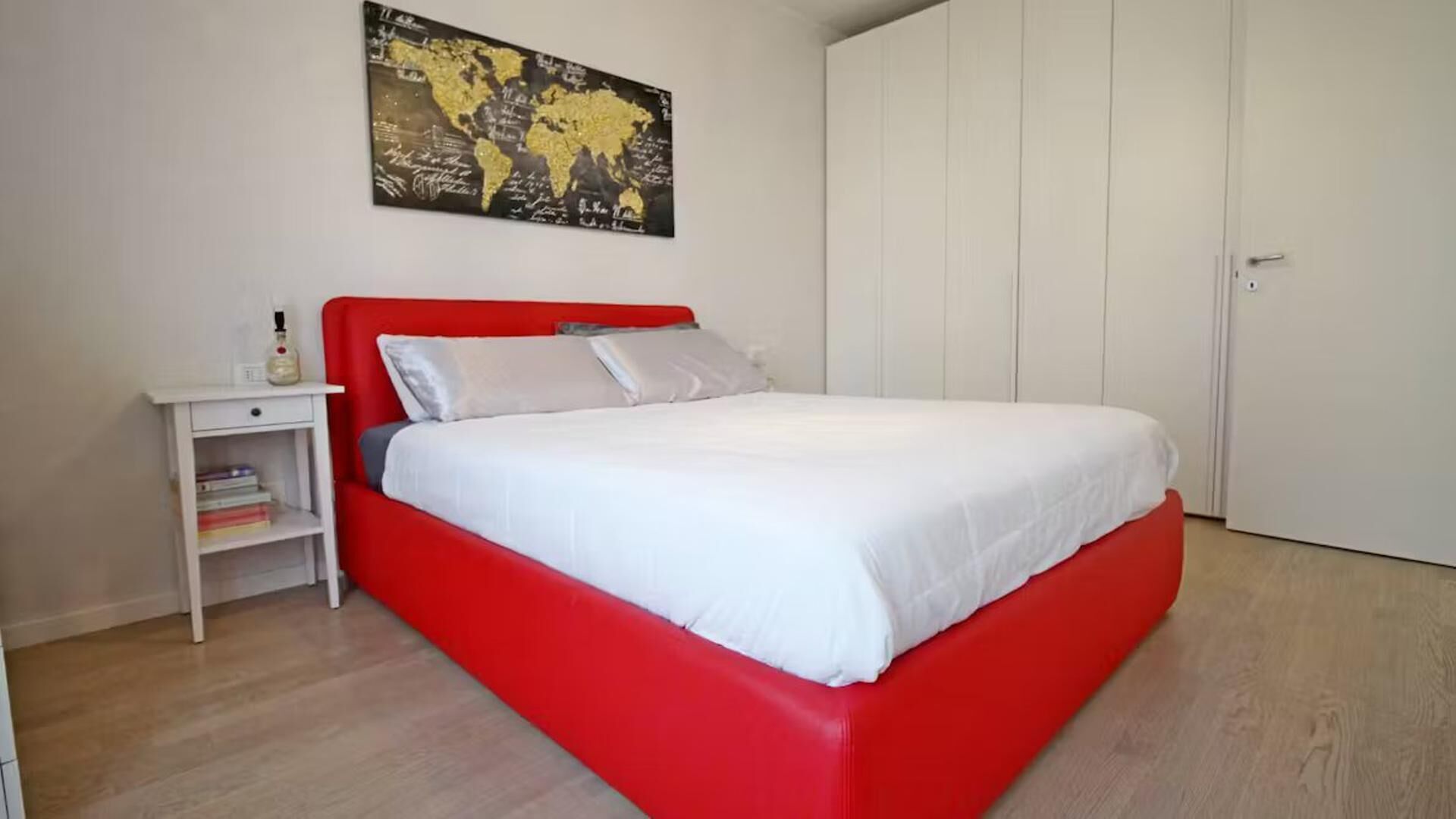 Une chambre avec un lit rouge, du linge de lit blanc, une table de chevet avec des livres et une carte du monde accrochée au mur.