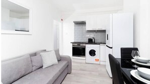 A modern apartment with a grey sofa, a dining area with a table and chairs, a kitchen with appliances, and a large window. - Flat 2 Covent Garden Apartments

 (London)