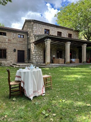 A stone building with a covered patio area, a table set for tea, and a grassy lawn.