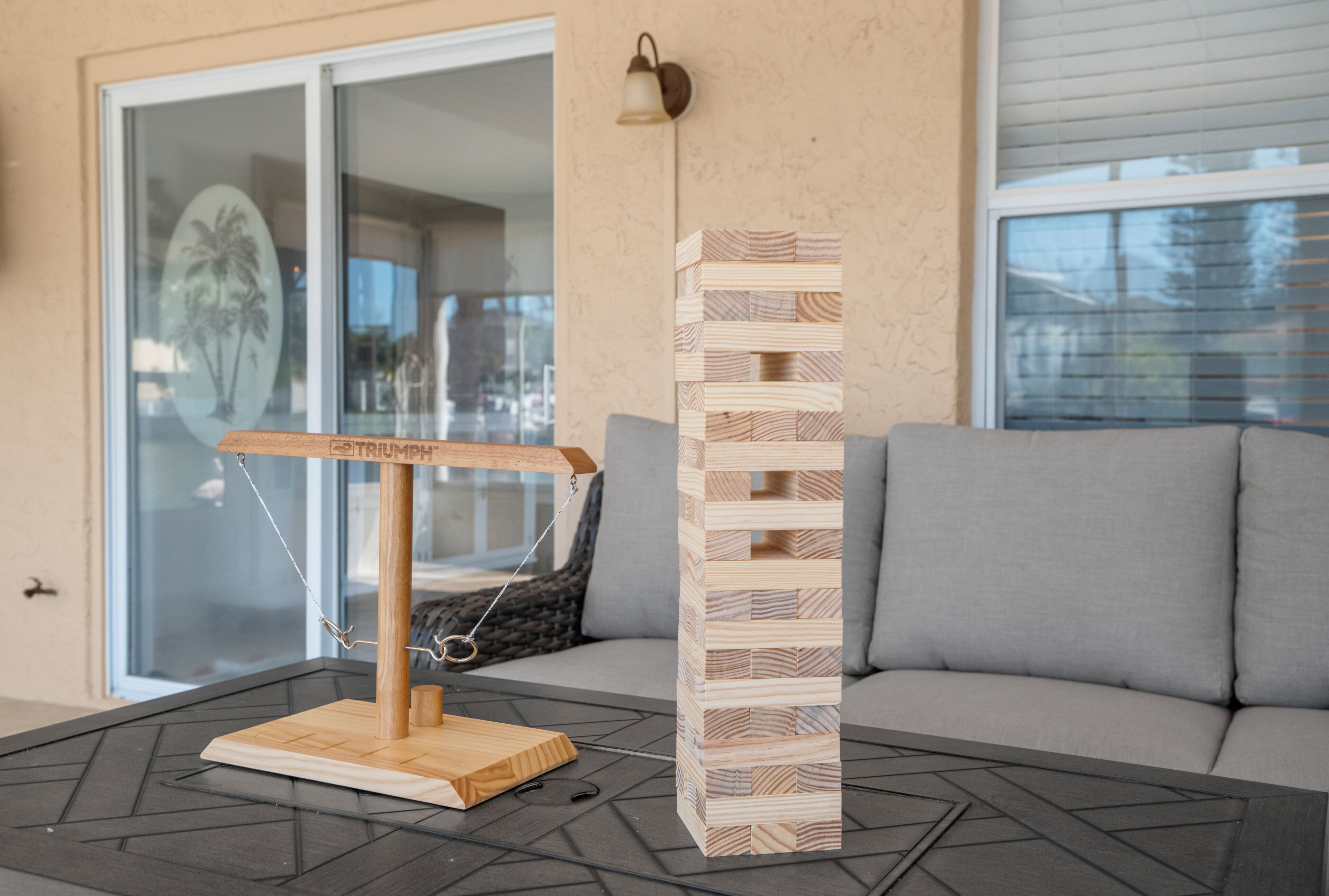Une tour de Jenga en bois, placée à côté d’une balançoire sur une table dans un salon, avec un canapé gris et une porte en verre en arrière-plan.