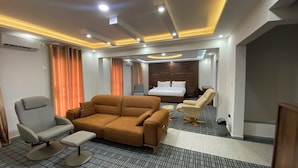 A hotel room with a large brown sofa, a grey armchair, a small table, and a bed with a wooden headboard. - LIA Hotel (Lungi)