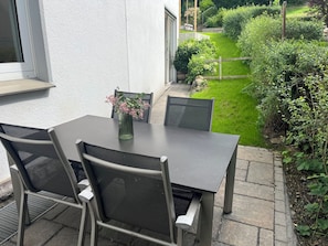 Un patio al aire libre con una mesa y sillas, un florero con flores y vistas a un jardín cuidado.