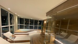 Una habitación de hotel moderna con un balcón acristalado, un jacuzzi y una zona de estar con sillones de descanso.