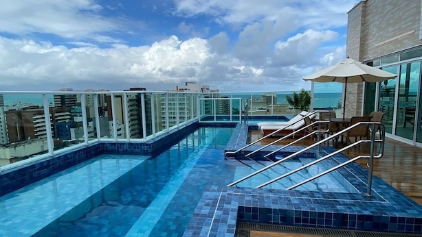 Una piscina infinita en la azotea con terraza de madera y sillones de descanso, con vistas a la ciudad y al océano.