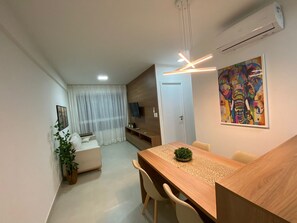 Een moderne woonkamer met een bank, een eettafel met stoelen, een aan de muur bevestigde televisie en een kleurrijk abstract schilderij.
