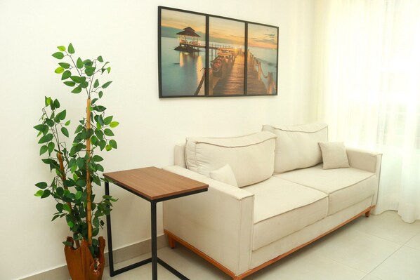 A living room with a beige sofa, a small wooden table, and a potted plant. - Edifício Promenade II Apt 303 (Maceió)