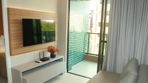 Una sala de estar moderna con un televisor de pantalla plana montado en un panel de madera, un mueble de entretenimiento blanco con un jarrón de flores y un ventanal corredizo que da a un balcón con un muro de azulejos verdes.