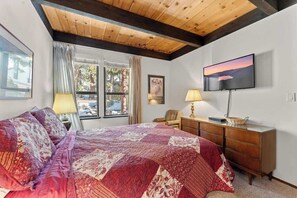 A bedroom with a large bed, a wooden ceiling, a TV, and a window with curtains.