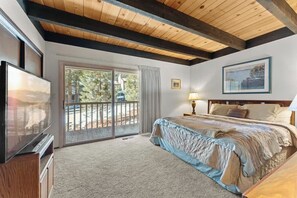 A bedroom with a large bed, a television, a balcony with a view of trees, and a framed picture on the wall.