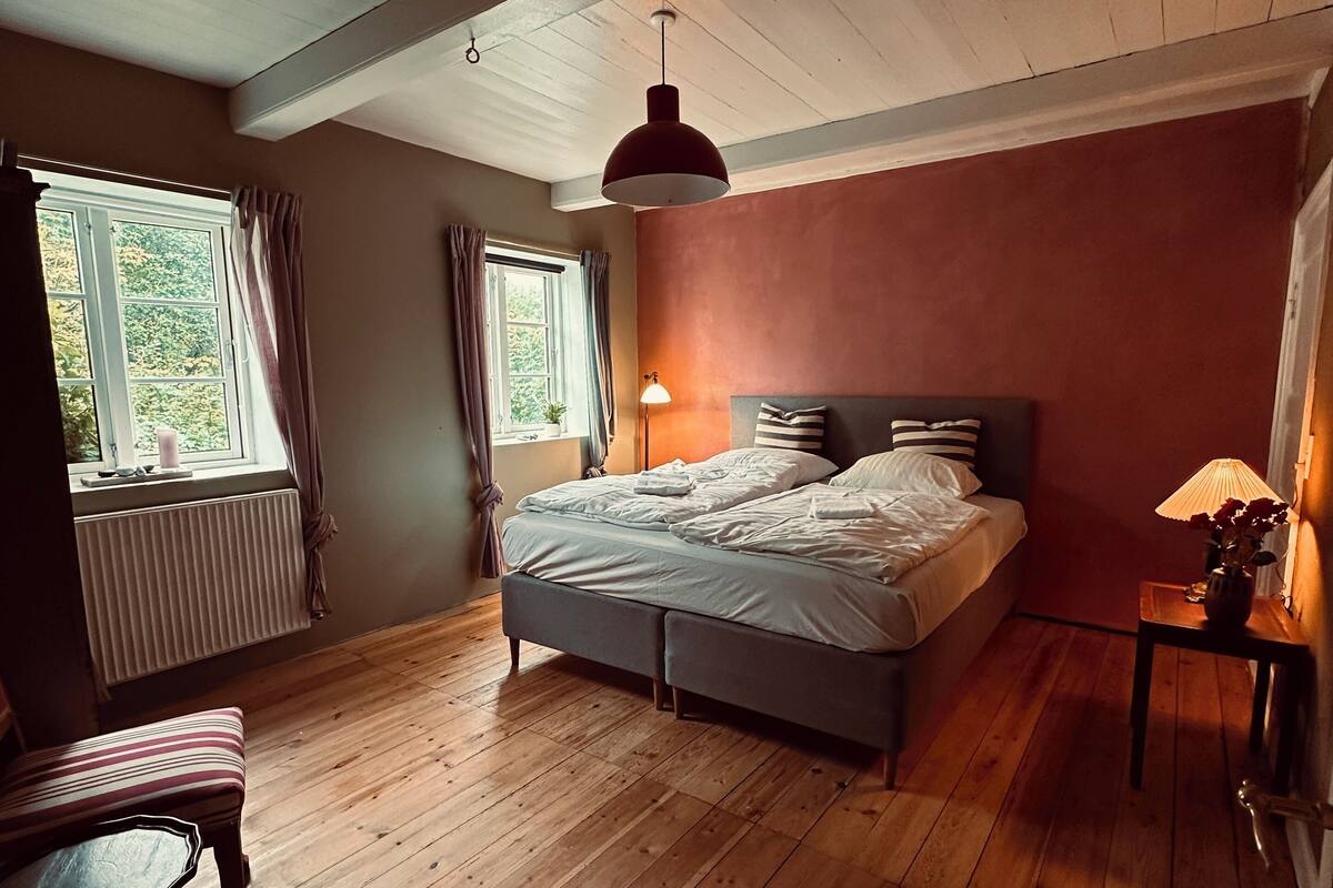 A bedroom with a large bed, wooden flooring, a window with curtains, and a ceiling light.