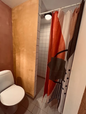 A bathroom with a white toilet, a shower with a red and white curtain, and tiled walls. - Bygagergaard (Askeby)