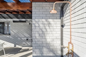 A modern outdoor shower with a copper showerhead, a brick wall, and a metal pipe system. - Sandy Shores Getaway (Mandurah)