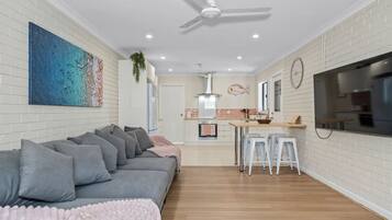 A modern living room with a grey sofa, a kitchen area with a TV, and a ceiling fan.