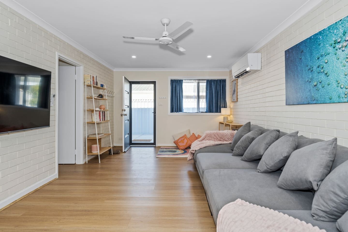 A modern living room with a grey sofa, a flat-screen TV, a ceiling fan, and a bookshelf.