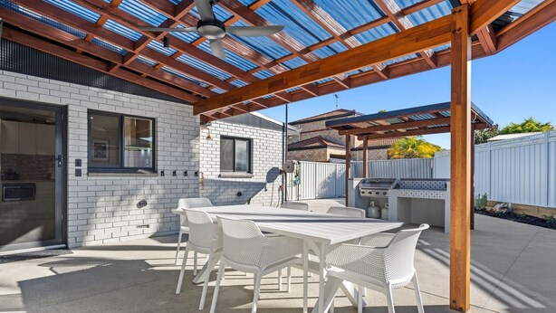 An outdoor patio with a covered seating area, a grill, and a white brick building in the background. - Sandy Shores Getaway (Mandurah)