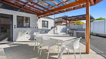 An outdoor patio with a covered seating area, a grill, and a white brick building in the background.