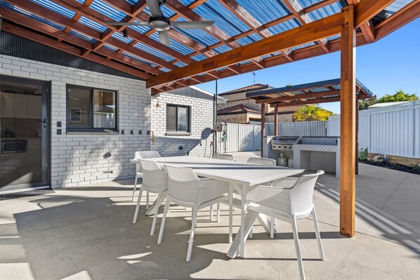 An outdoor patio with a covered seating area, a grill, and a white brick building in the background.