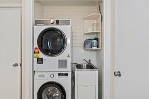 A compact laundry room with stacked washing machines, a sink, and a shelf with toiletries.