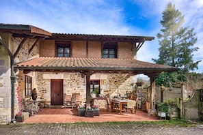 A stone house with a tiled roof, a covered patio with furniture, and a wooden fence.