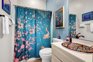 A bathroom with a blue shower curtain featuring cherry blossoms, a white toilet, a sink with a colorful basin, and two framed artworks on the wall.