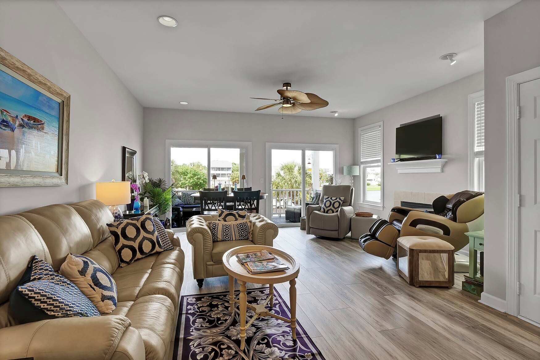 A spacious living room with a leather sofa, armchairs, a coffee table, and a ceiling fan. A dining area with chairs and a table is visible through the glass doors.