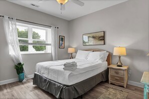 A bedroom with a bed, two bedside tables with lamps, a window with curtains, a potted plant, and a painting on the wall.