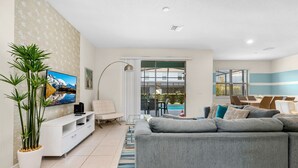 A modern living room with a grey sofa, a white entertainment unit, a large flat-screen TV, and a potted plant. - Family home w/ private pool near Disney (Kissimmee)