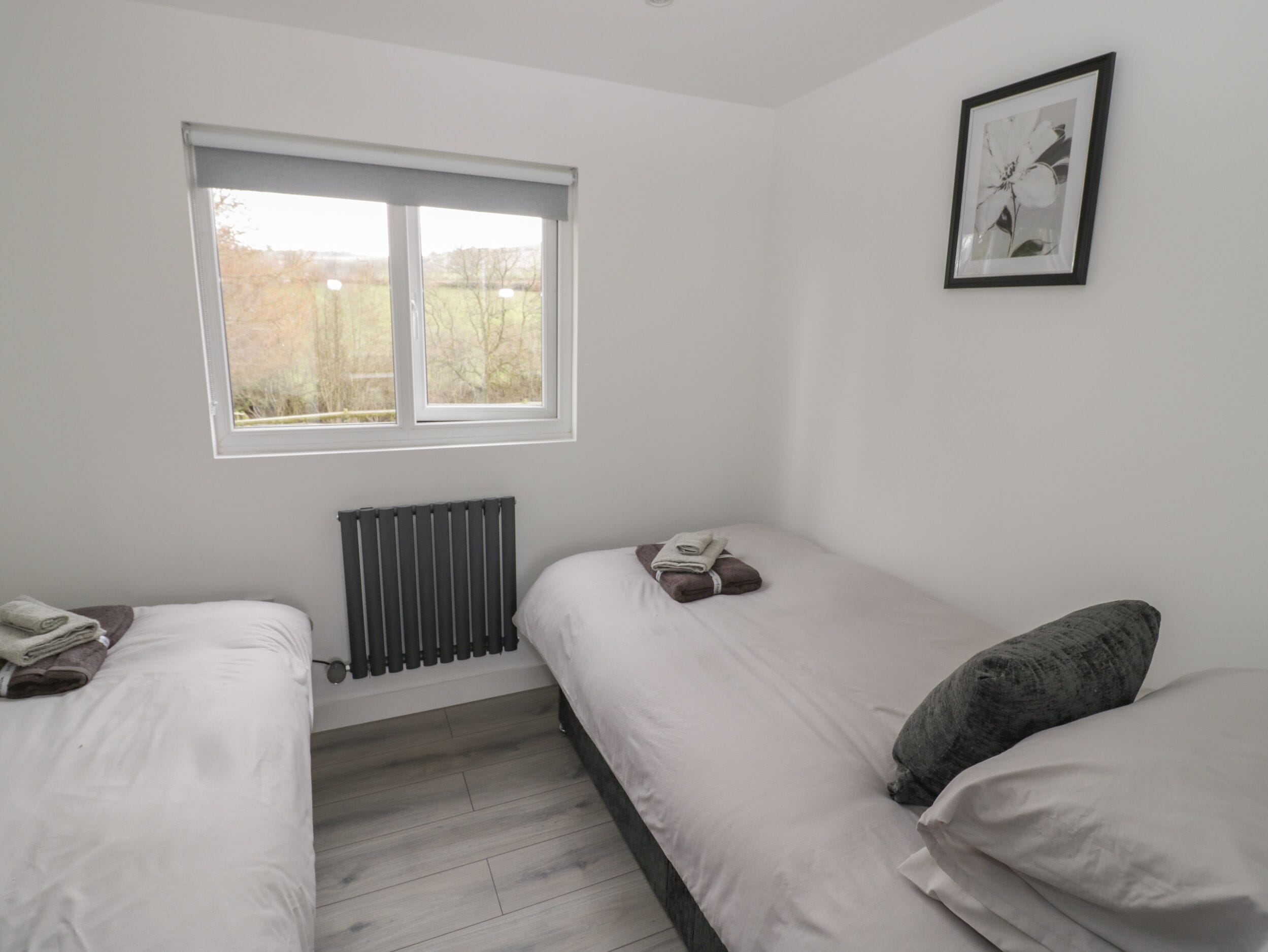 A bedroom with two single beds, a radiator, a window with a view of trees, and a framed picture on the wall.