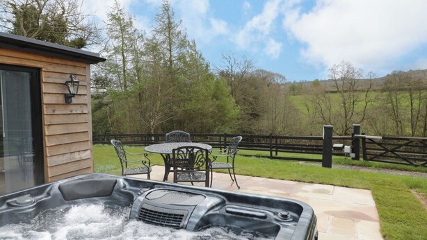 Ein Whirlpool mit Holzterrasse und Gartenmöbeln, umgeben von einem Holzzaun und Bäumen.