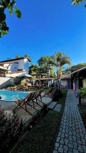 A pool area with lounge chairs, a paved walkway, and a building with a tiled roof. - Pousada Tropicália (Porto Seguro)