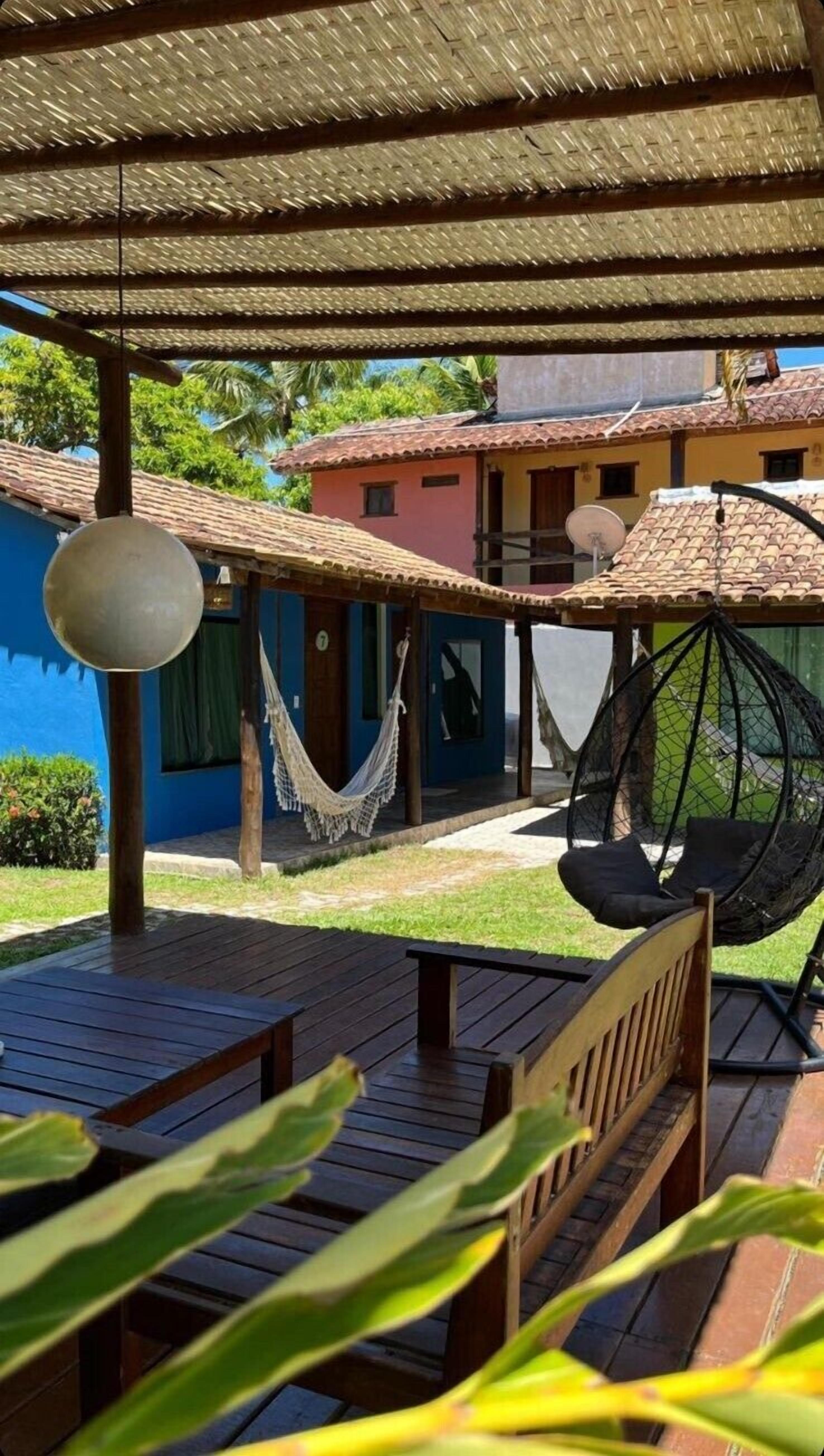 Una terraza de madera con una hamaca y una silla colgante, un edificio azul al fondo y una estructura de techo de paja.